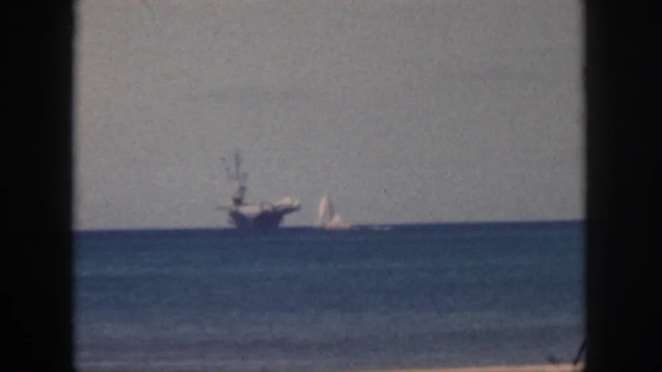 1959 Oahu home-movie frame with a distant aircraft carrier, white sailboat, calm water, muted sky, and dark window framing at both sides.