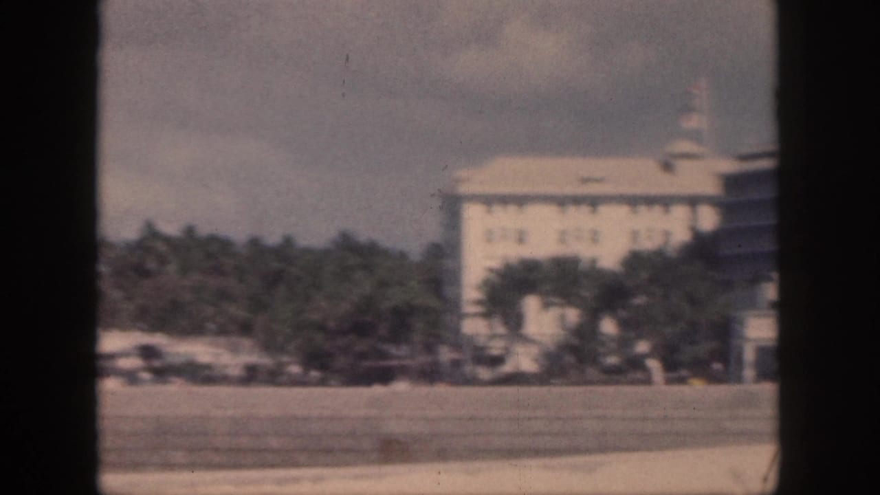 1959 Oahu frame with a large pale waterfront building, palms, and shoreline foreground in soft 8mm color.