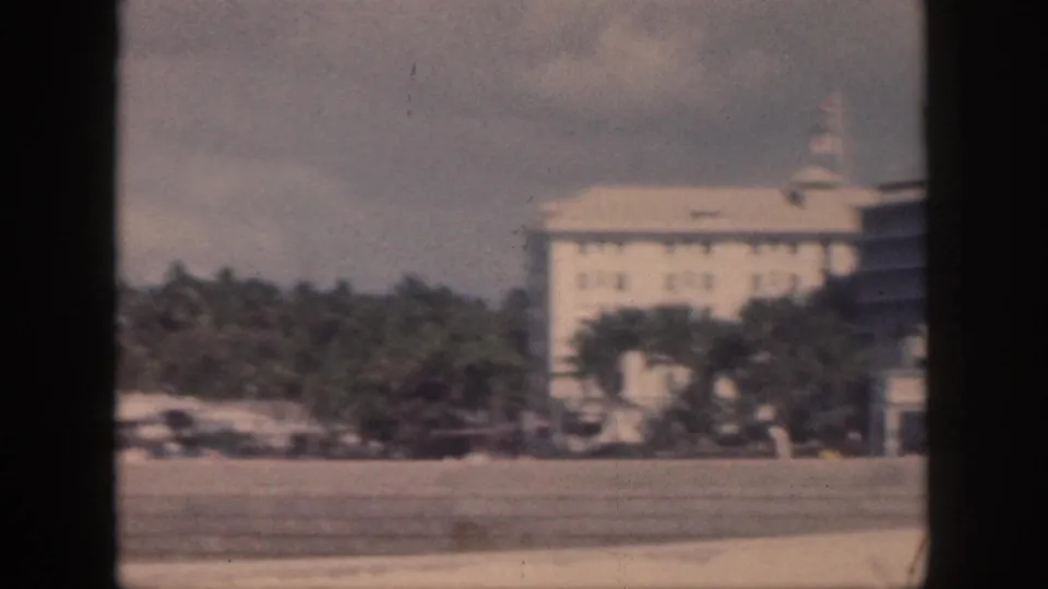 1959 Oahu frame with a large pale waterfront building, palms, and shoreline foreground in soft 8mm color.