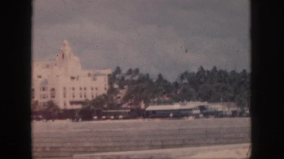 1959 Oahu home-movie frame showing a large pale coastal building, palms, and vehicles along the waterfront.