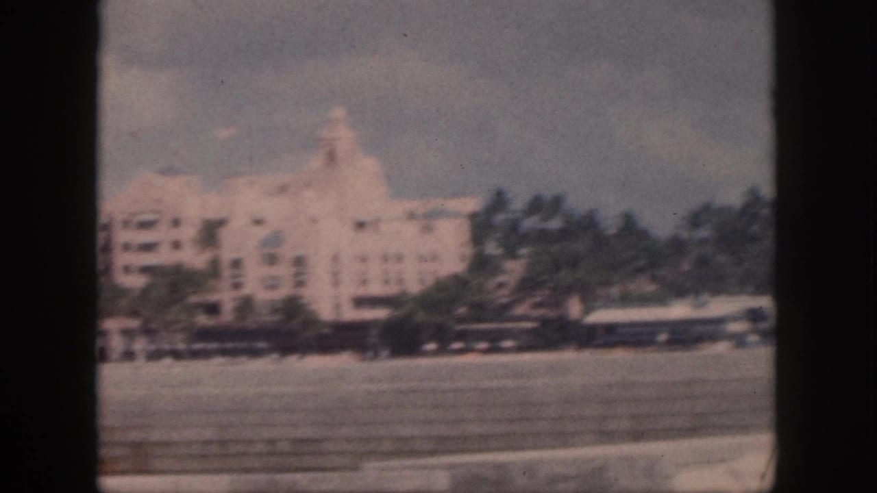 1959 Oahu 8mm frame showing a large pale hotel-like building, palm trees, and a paved foreground from a moving vehicle.