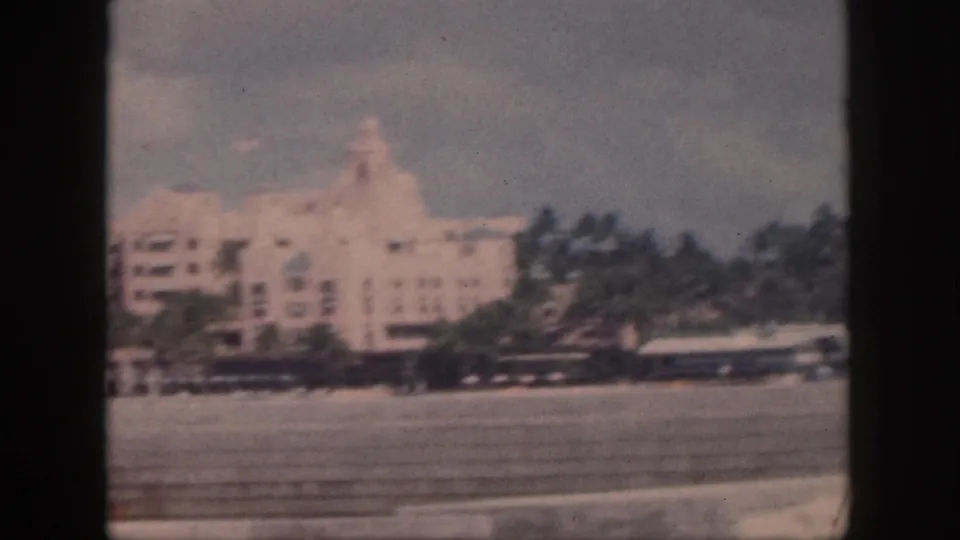 1959 Oahu 8mm frame showing a large pale hotel-like building, palm trees, and a paved foreground from a moving vehicle.