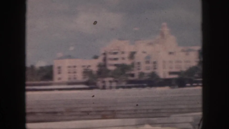 1959 Oahu home-movie frame of a large white stepped building and palms, viewed from a moving vehicle.
