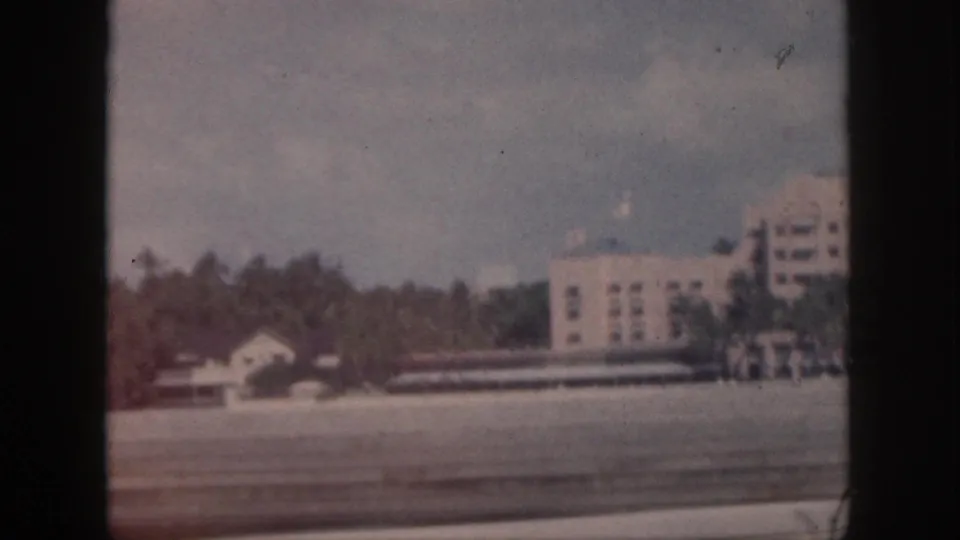 1959 Oahu frame with the building partly cropped, palms, a smaller structure, and roadside foreground blur.