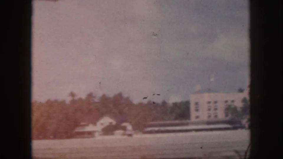 1959 Oahu 8mm frame showing a small roadside structure, tree line, and the edge of a larger white building.