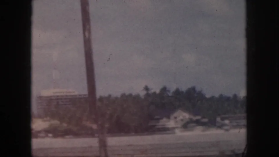 1959 Oahu shoreline with palms and distant buildings, viewed through a dark window or structural frame on 8mm film.