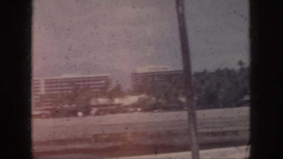 Oahu waterfront scene with low buildings, palms, and a large distant structure, partially obscured by window framing.