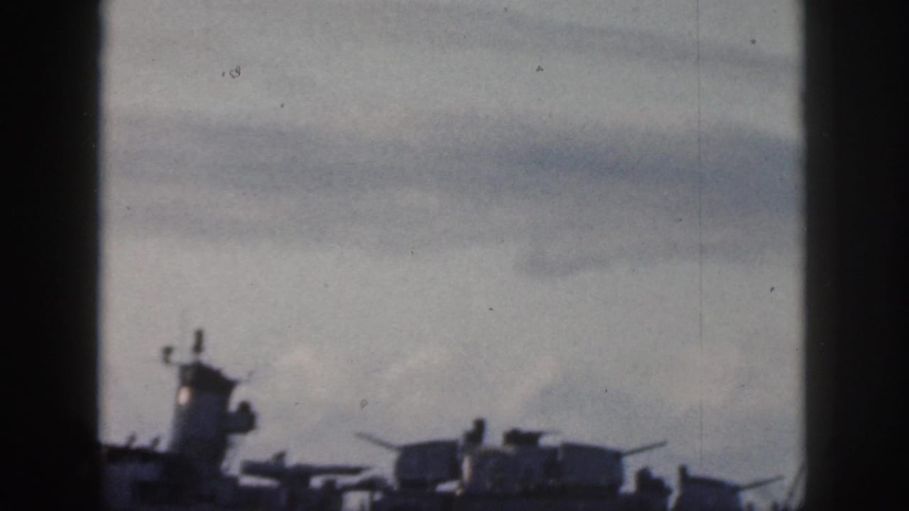 Silhouetted naval ship with turrets under a cloudy sky.