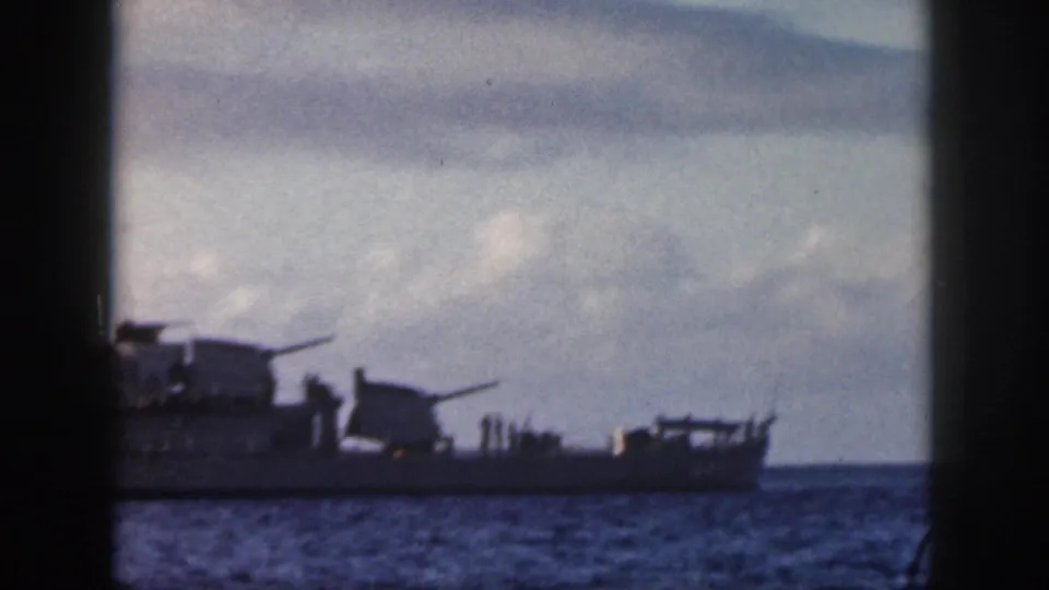1959 Oahu home-movie frame of a naval ship at sea, viewed through a dark vertical foreground frame with cloudy sky behind it.