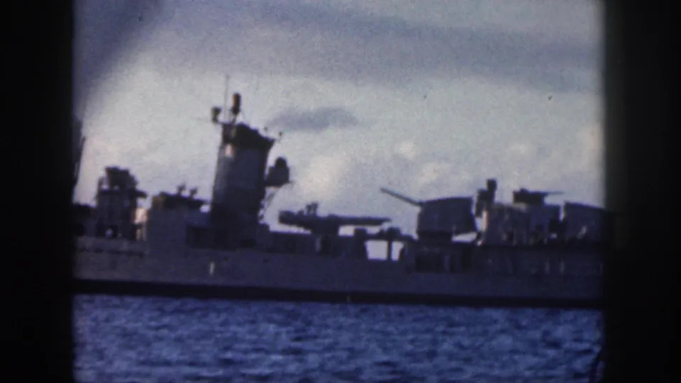 1959 Oahu 8mm frame showing the side of a Navy warship with superstructure, gun mounts, and railings over calm sea.