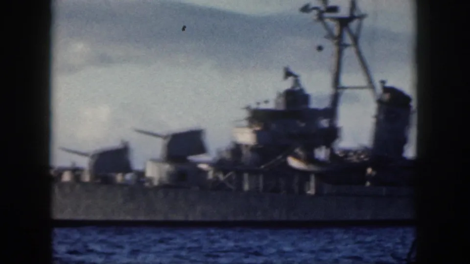 Gray naval vessel near Oahu with radar mast, superstructure, gun mounts, and railings visible above the ocean in grainy 8mm footage.