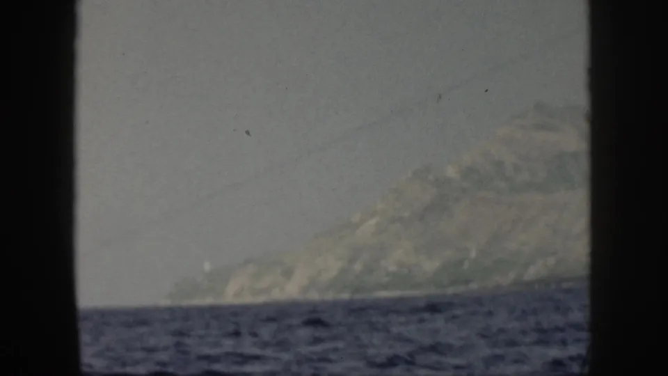 Oahu coastline in a 1959 8mm frame, with a hazy mountain slope rising beyond dark ocean water.