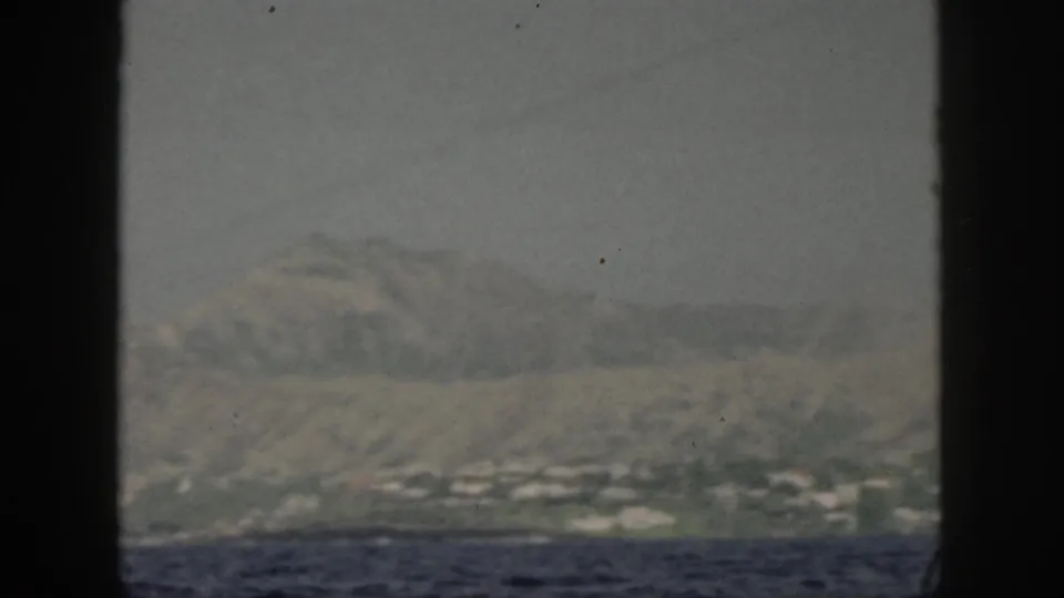 A distant Oahu shoreline with pale buildings below rugged slopes is seen from a moving boat in muted 8mm color.