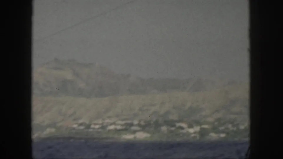 The boat view holds on a hazy Oahu coast, with scattered light buildings at the base of broad mountain slopes.