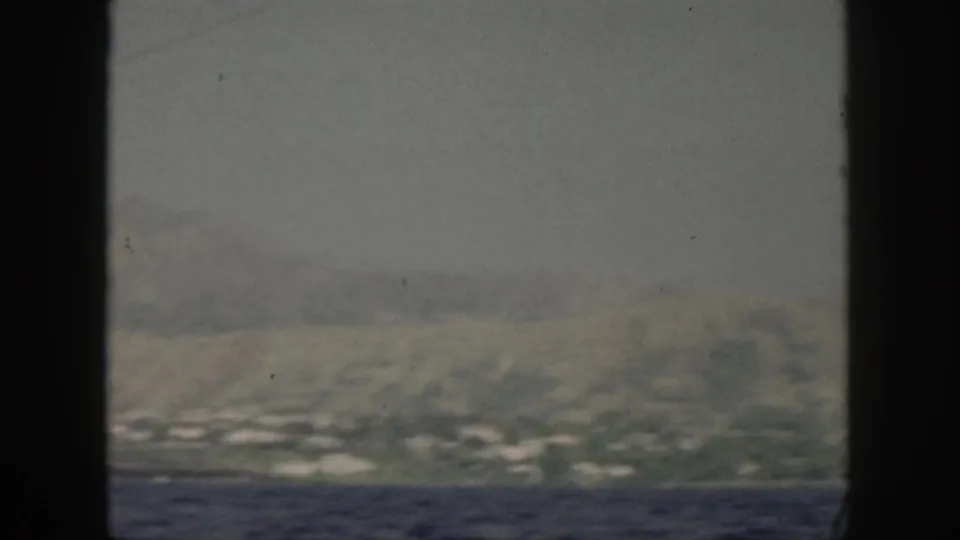A muted 8mm frame shows the Oahu shoreline receding in haze, with ocean in front and mountain slopes beyond.