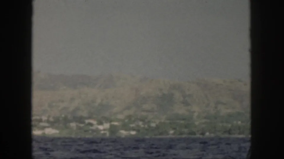 1959 Oahu coastline viewed from a boat, with ocean foreground, hazy hills, pale buildings, and dark window framing.