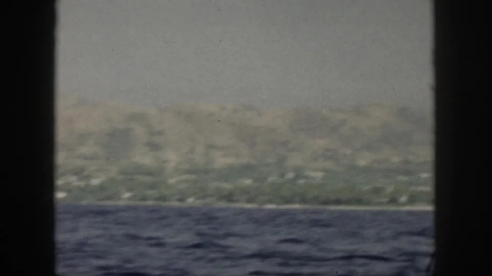 Hazy 1959 Oahu shoreline from a moving boat, showing ocean, low coast, green hills, and soft home-movie color.