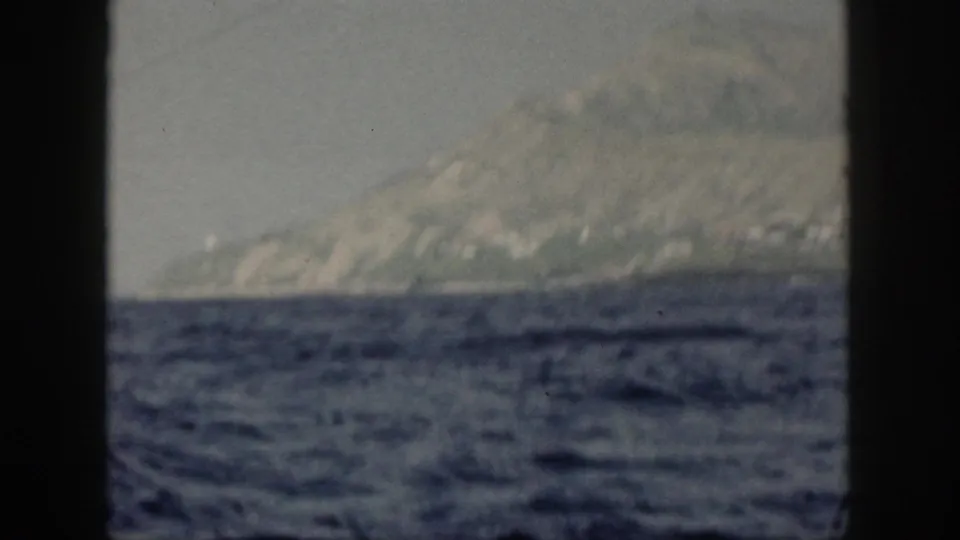1959 Oahu coastal hills and mountain slope seen across choppy ocean, with a dark boat-window divider at the left edge.
