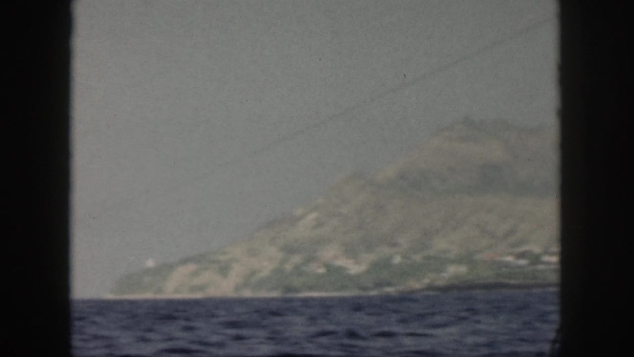 1959 Oahu home-movie frame showing a hazy mountain coastline across choppy water, viewed through a dark boat or window frame.