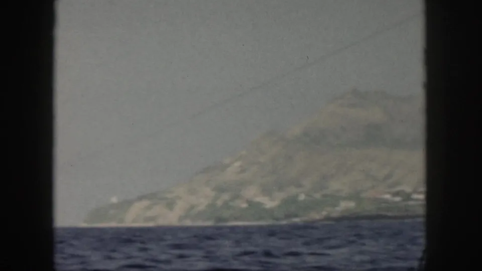 1959 Oahu home-movie frame showing a hazy mountain coastline across choppy water, viewed through a dark boat or window frame.