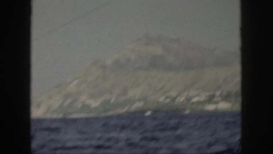 Distant Oahu coastline in 1959, with muted green slopes above choppy water and a dark vertical frame edge in the foreground.