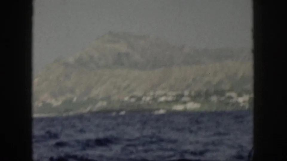 1959 Oahu coastal view with small light shoreline structures beneath a hazy mountain ridge, filmed from a moving boat.