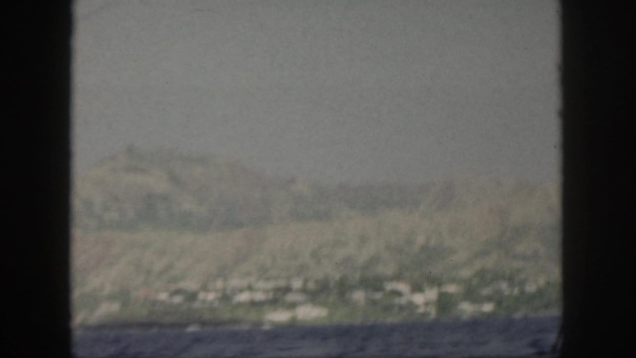 1959 Oahu coastline seen from a moving boat, with hazy hills, shoreline greenery, and dark water framed by black film edges.
