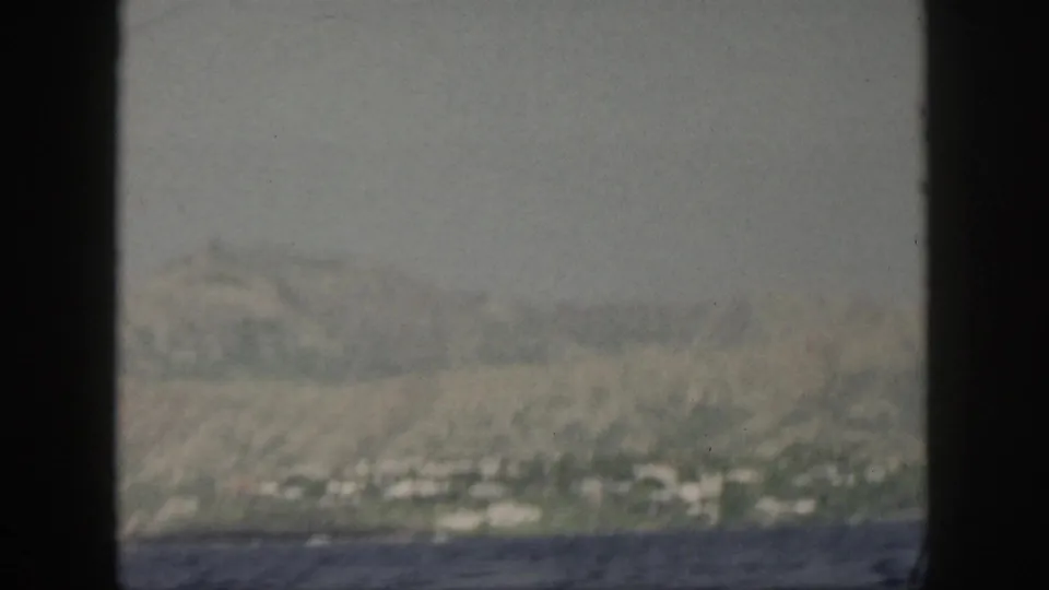 1959 Oahu coastline seen from a moving boat, with hazy hills, shoreline greenery, and dark water framed by black film edges.
