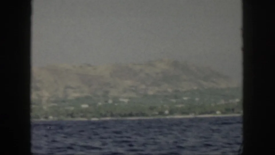 1959 Oahu shore viewed from offshore, with green hills, a narrow coastline, scattered buildings, and heavy dark film-gate edges.