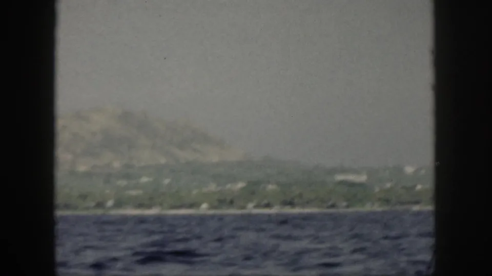 Soft 1959 home-movie view of Oahu shoreline from the water, showing greenery, low hills, and a hazy sky through black frame edges.