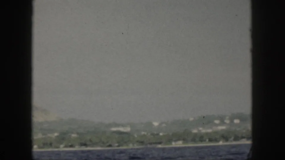 1959 Oahu coastal hillside seen from a boat, with pale buildings scattered above the shore and muted film color.