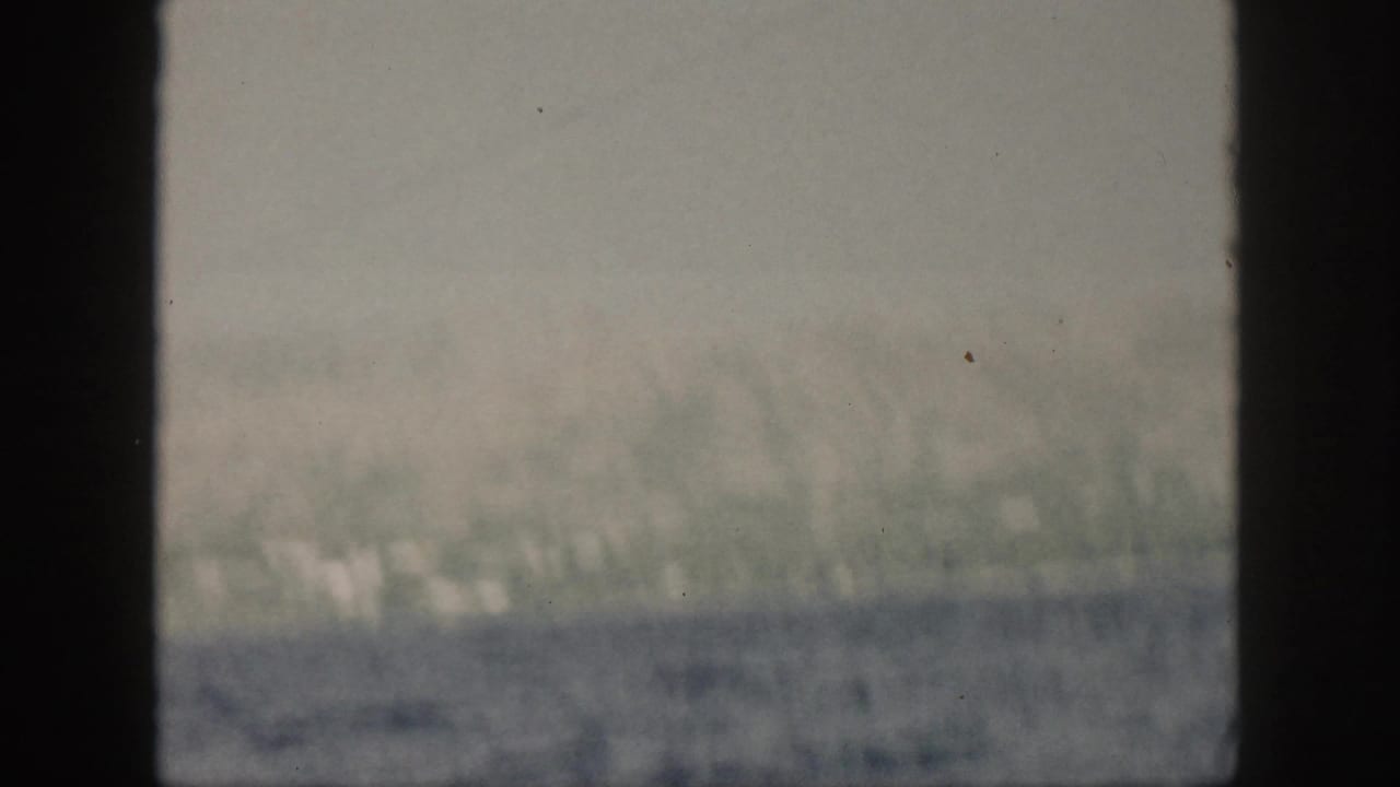 1959 Oahu coastal view through a window frame, with hazy green hills, pale shoreline structures, and blue ocean in the foreground.
