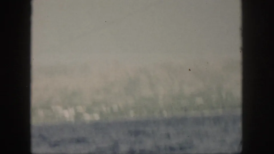 1959 Oahu coastal view through a window frame, with hazy green hills, pale shoreline structures, and blue ocean in the foreground.