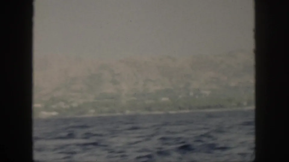 Through a dark window frame, the 1959 Oahu view shows rippled blue water, a hazy shoreline, and soft green hills beyond.