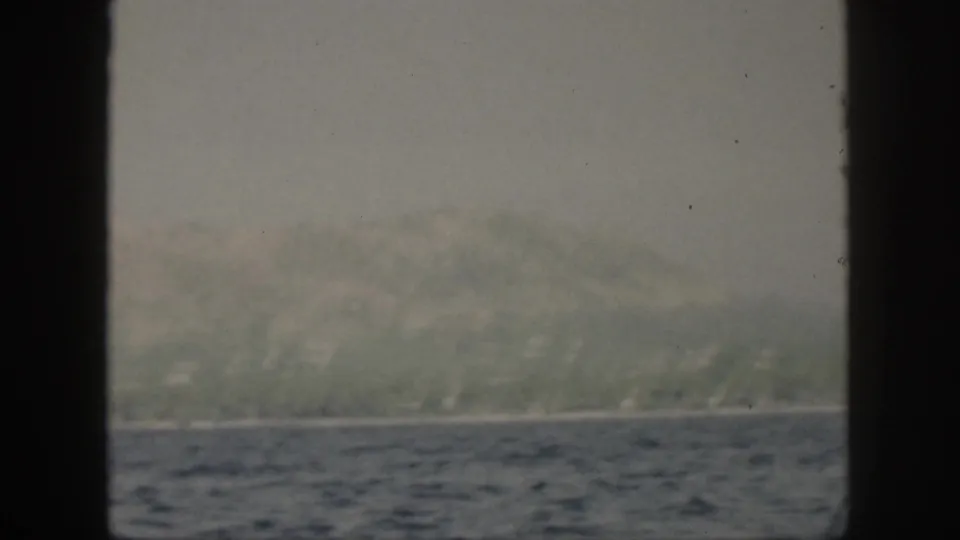 A soft 8mm view of Oahu in 1959 shows distant green hills, a narrow coast, and blue ocean from behind a window frame.