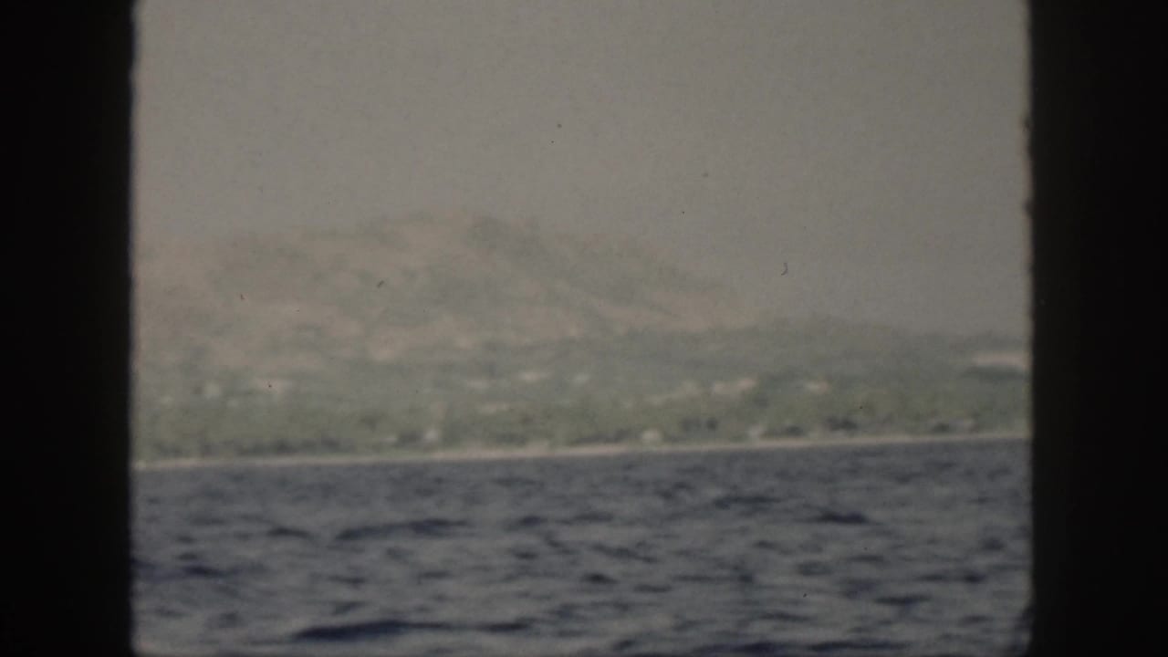 1959 Oahu home-movie frame showing a hazy mountain coast beyond blue ocean water, viewed from a boat through dark framing.