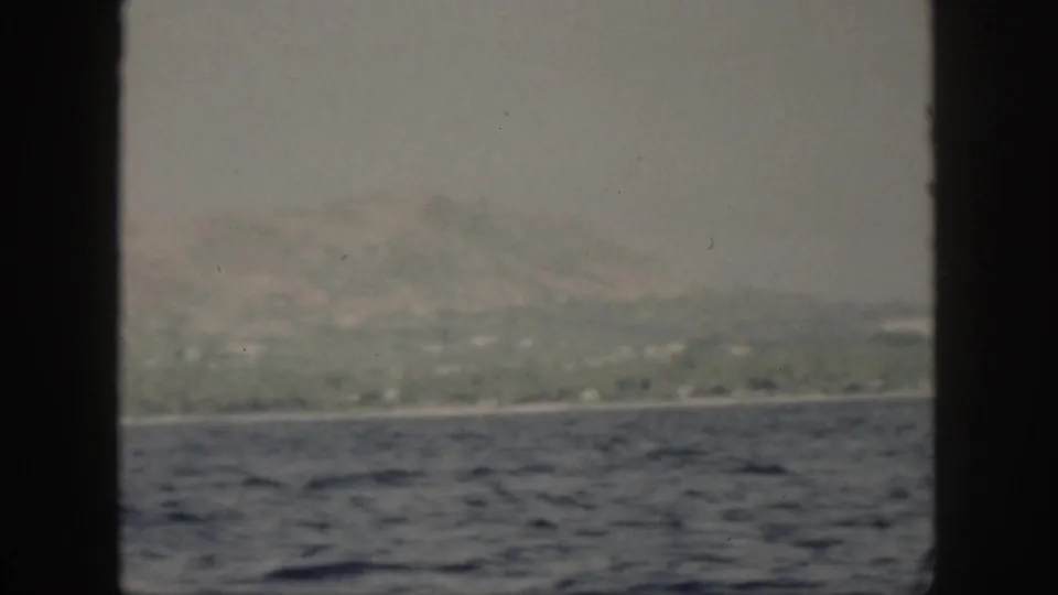1959 Oahu home-movie frame showing a hazy mountain coast beyond blue ocean water, viewed from a boat through dark framing.