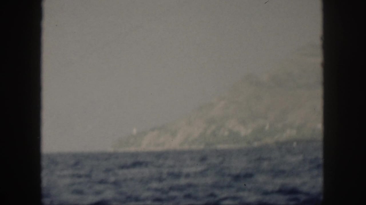 1959 Oahu coastal view with a mountain slope and shoreline across blue water, partially obscured by a dark window or frame edge.