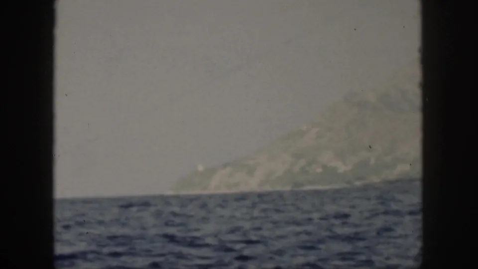 1959 Oahu home-movie still of a greenish mountain coastline seen across ocean water from a moving boat.