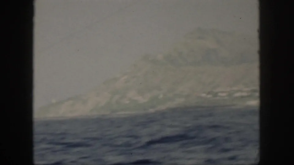 1959 Oahu footage frame showing the island coastline through haze, with ocean in the foreground and dark framing at the side.