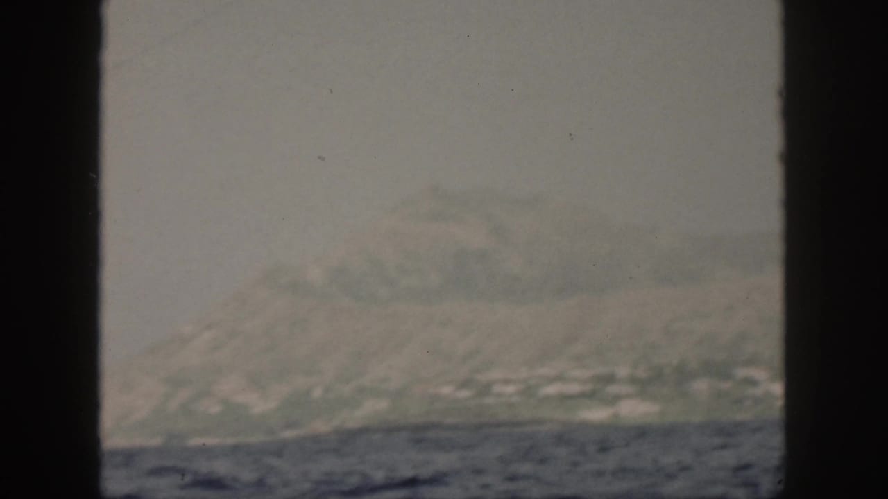 1959 Oahu home-movie frame showing a hazy island shoreline beyond choppy water, partly blocked by dark frame edges.