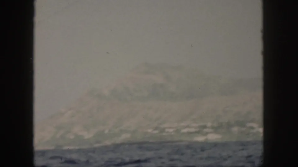 1959 Oahu home-movie frame showing a hazy island shoreline beyond choppy water, partly blocked by dark frame edges.