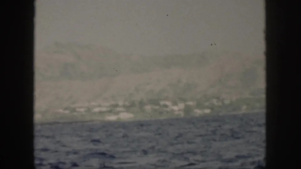 1959 Oahu home-movie frame showing misty cliffs, scattered pale shoreline structures, and rough water from offshore.