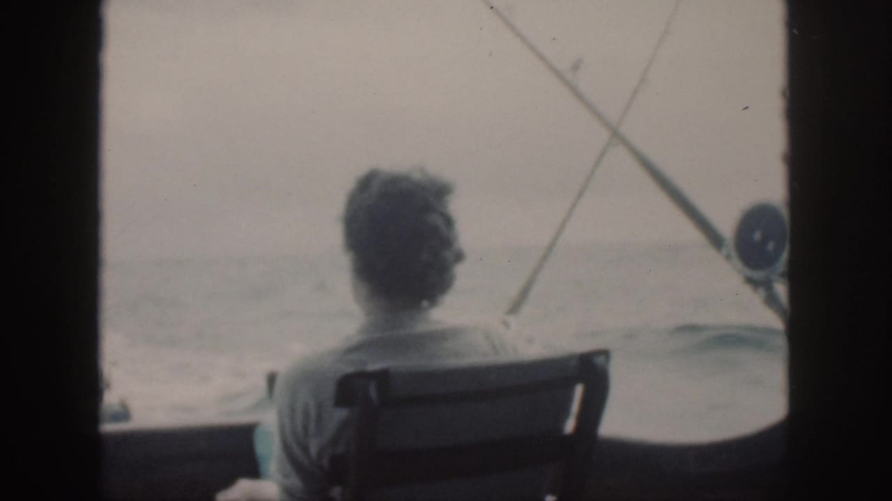 Man seated on a boat facing the sea with fishing rods nearby.