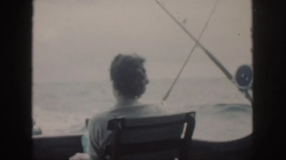 A man sits in a wooden chair on a boat off Oahu, facing the sea with fishing rods visible beside him.