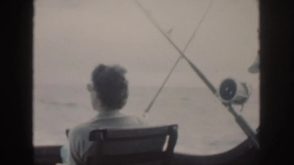 Rear view of the seated man in the 1959 Oahu boat scene, with fishing rods and a reel visible against the sea.