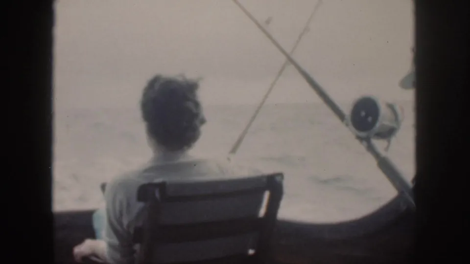 A woman sits with her back to the camera on a 1959 Oahu fishing boat, framed by rods, reel, and pale sea beyond.