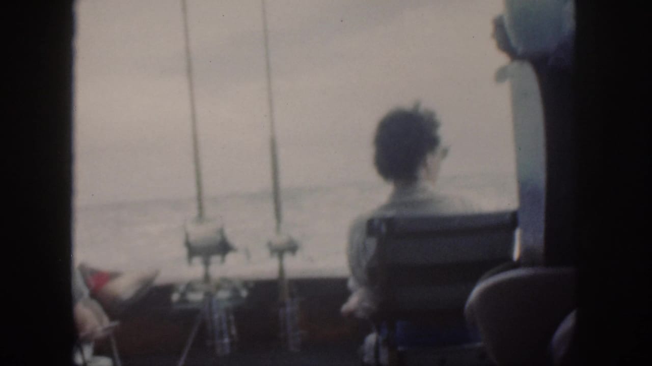 A seated figure is seen from behind on a 1959 Oahu fishing boat, with rods mounted near the rail and open water beyond.