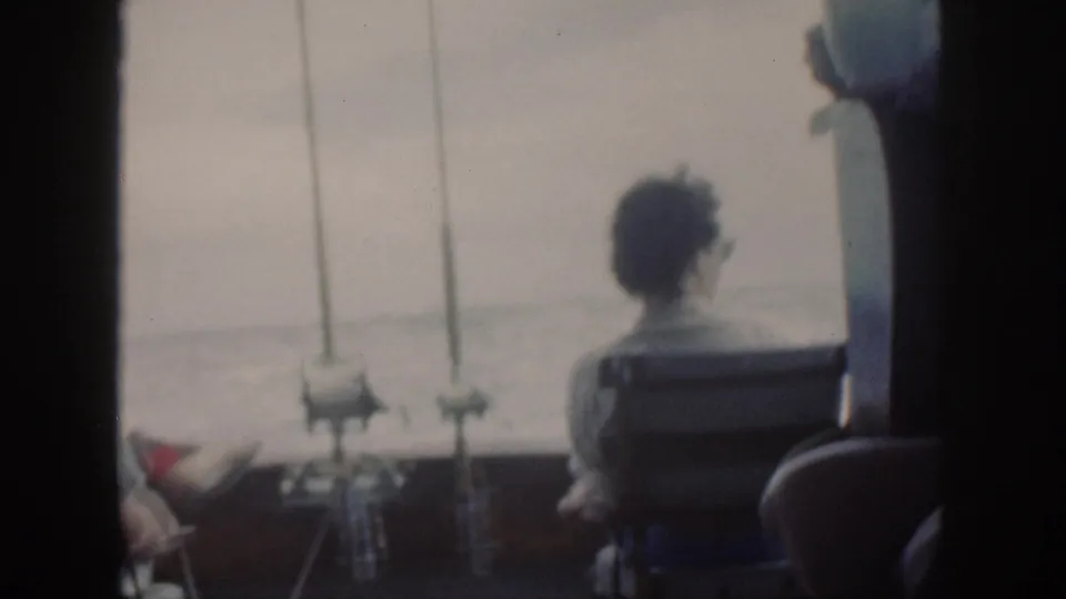A seated figure is seen from behind on a 1959 Oahu fishing boat, with rods mounted near the rail and open water beyond.