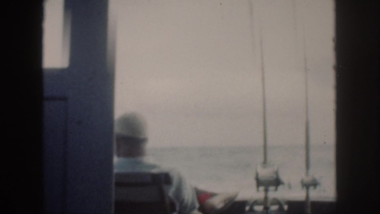 1959 Oahu home-movie frame showing a seated adult in a cap on a boat, viewed through dark interior framing toward the sea.